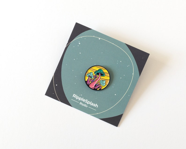 May include: A colorful enamel pin featuring a cluster of pink and purple mushrooms with green tops, set against a yellow and orange background. The pin is on a card with the text "RippleSplash Studio".