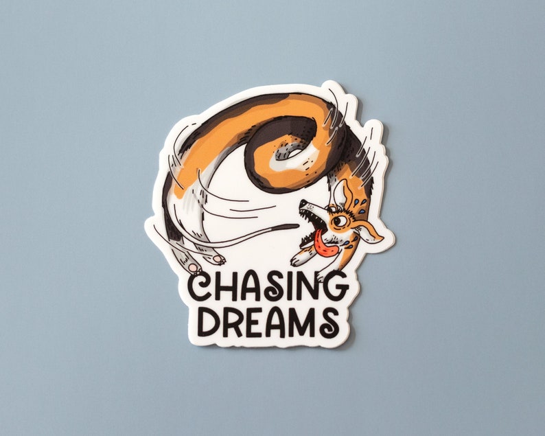 May include: A white sticker with a cartoon illustration of a corgi dog chasing its tail. The dog is brown, black, and white, with its tongue sticking out. The words "CHASING DREAMS" are in black, bold letters.