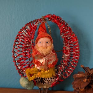 May include: A vintage Christmas ornament featuring a gnome figure reading a book with "NOEL" on the cover. The gnome wears a red hat and has a white beard. The ornament is set within a red wire frame.