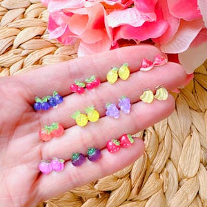 May include: A set of 10 pairs of fruit-shaped earrings. The earrings are made of clear plastic and feature a variety of fruits, including strawberries, cherries, grapes, lemons, pineapples, and bananas.