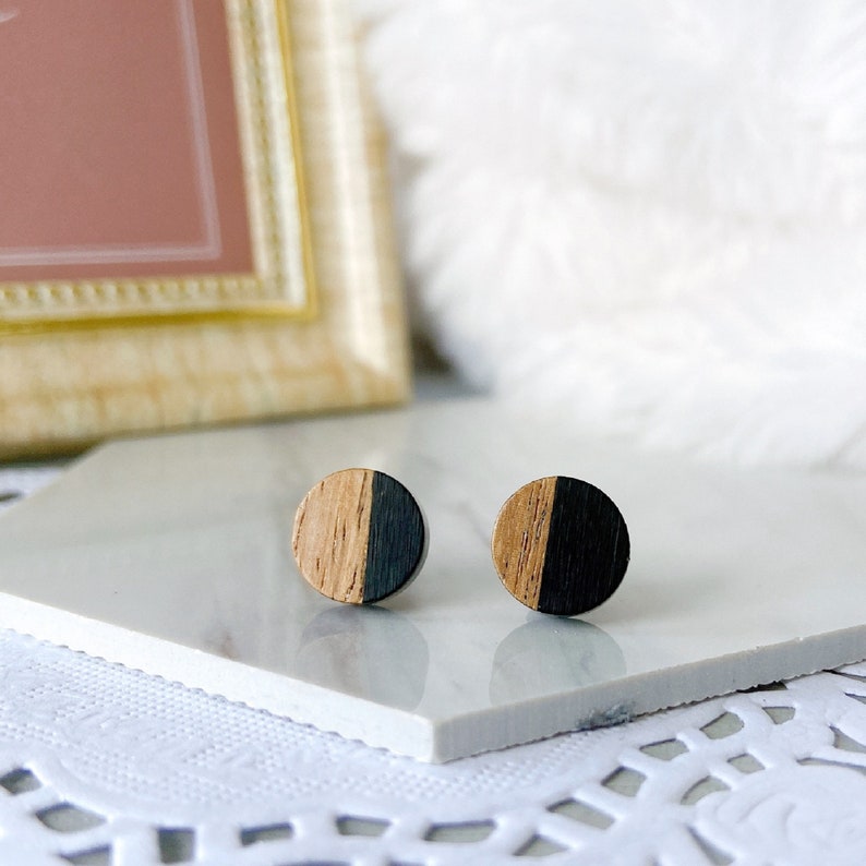 May include: A pair of round wooden stud earrings. Each earring is split into two halves: one half is natural wood grain, and the other is black. The earrings are displayed on a white surface.