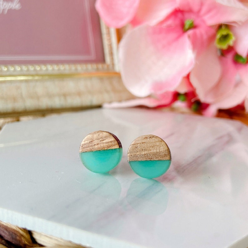 May include: A pair of round stud earrings. Each earring features a half-circle of light wood and a half-circle of turquoise resin. The earrings are displayed on a white surface with a blurred pink floral background.