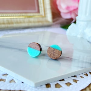 May include: A pair of round stud earrings. Each earring is split diagonally, with a section of dark wood and a section of bright turquoise. The earrings are displayed on a white marble surface.