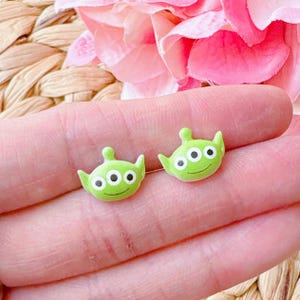 Op de afbeelding: Een paar groene alien oorbel studs met drie ogen en een glimlachend gezicht. De oorbellen zijn gemaakt van polymer clay en zijn ongeveer 1 cm in diameter.