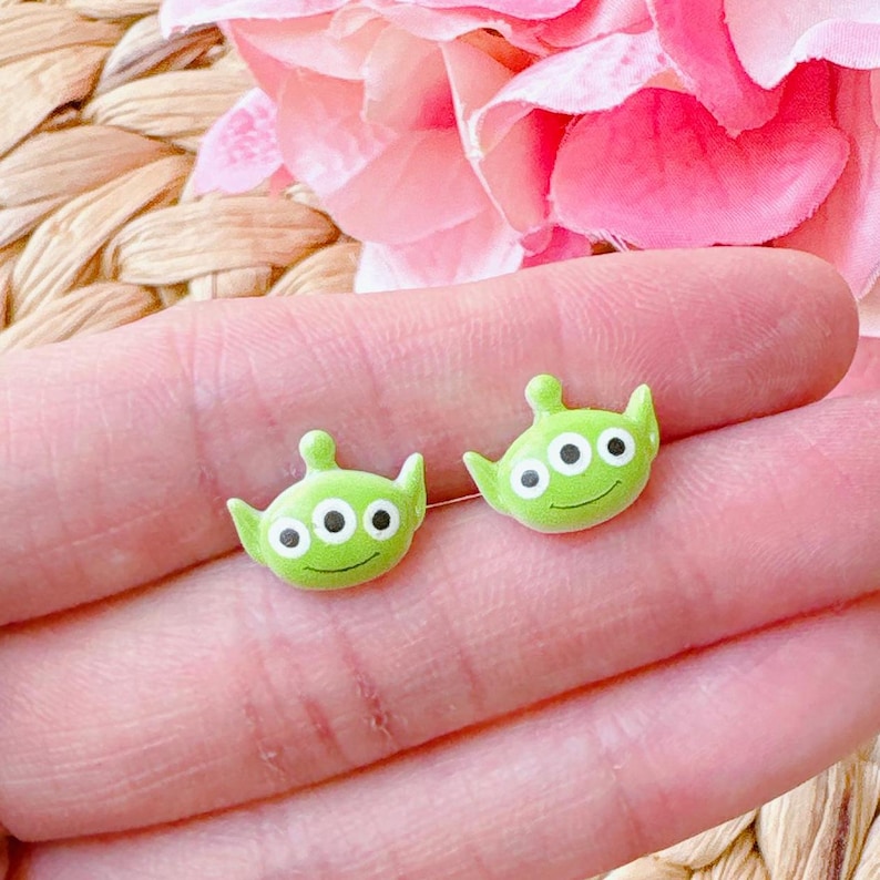 Op de afbeelding: Een paar groene alien oorbel studs met drie ogen en een glimlachend gezicht. De oorbellen zijn gemaakt van polymer clay en zijn ongeveer 1 cm in diameter.