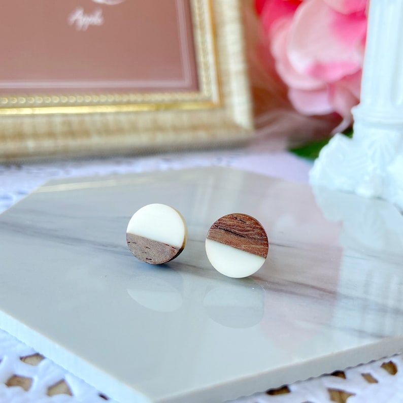 May include: A pair of round stud earrings. Each earring features a half-circle of light wood grain and a half-circle of white resin. The earrings are displayed on a white and gray marble surface.