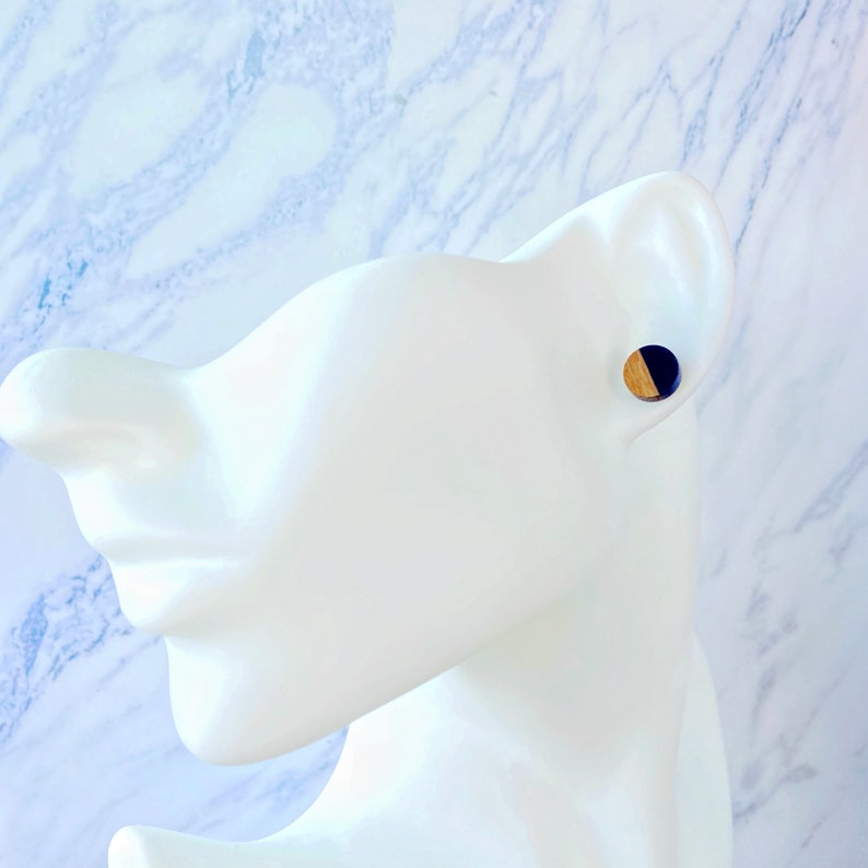 May include: A pair of round stud earrings. Each earring features a half-circle of natural wood and a half-circle of dark blue material. The earrings are displayed on a white mannequin head against a marble background.