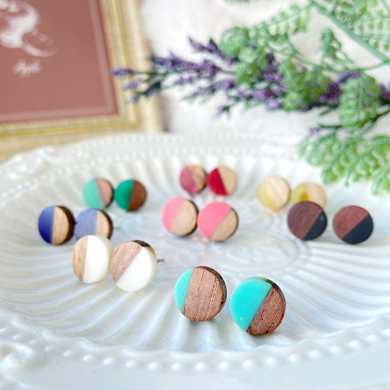 May include: A collection of colorful stud earrings displayed on a white, patterned plate. Each earring features a wooden half-circle paired with a solid color or white half-circle. Colors include blue, pink, green, and brown.