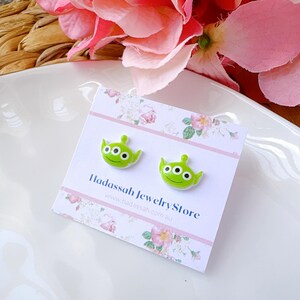 Op de afbeelding: Een paar groene alien-vormige oorbel studs op een witte kaart met de tekst "Hadasssah Jewelry Store" en het website adres "www.hadasssah.com.au".