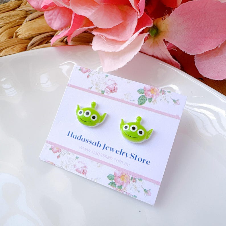 Op de afbeelding: Een paar groene alien-vormige oorbel studs op een witte kaart met de tekst "Hadasssah Jewelry Store" en het website adres "www.hadasssah.com.au".