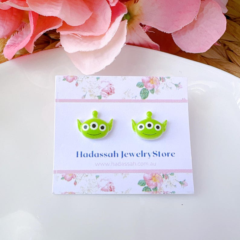 Op de afbeelding: Een paar groene alien oorbel studs met elk drie ogen. De oorbellen zijn op een witte kaart met roze bloemen en de tekst "Hadasssah Jewelry Store www.hadasssah.com.au".