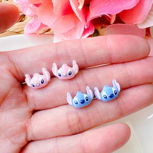 May include: Four small, colourful, cartoon character earrings. Two are pink with black eyes and two are blue with black eyes.