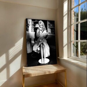 May include: Black and white framed print of a woman in a robe, seated and holding a phone and a book. The print is displayed on a woven bench with gold legs, near a window with natural light.