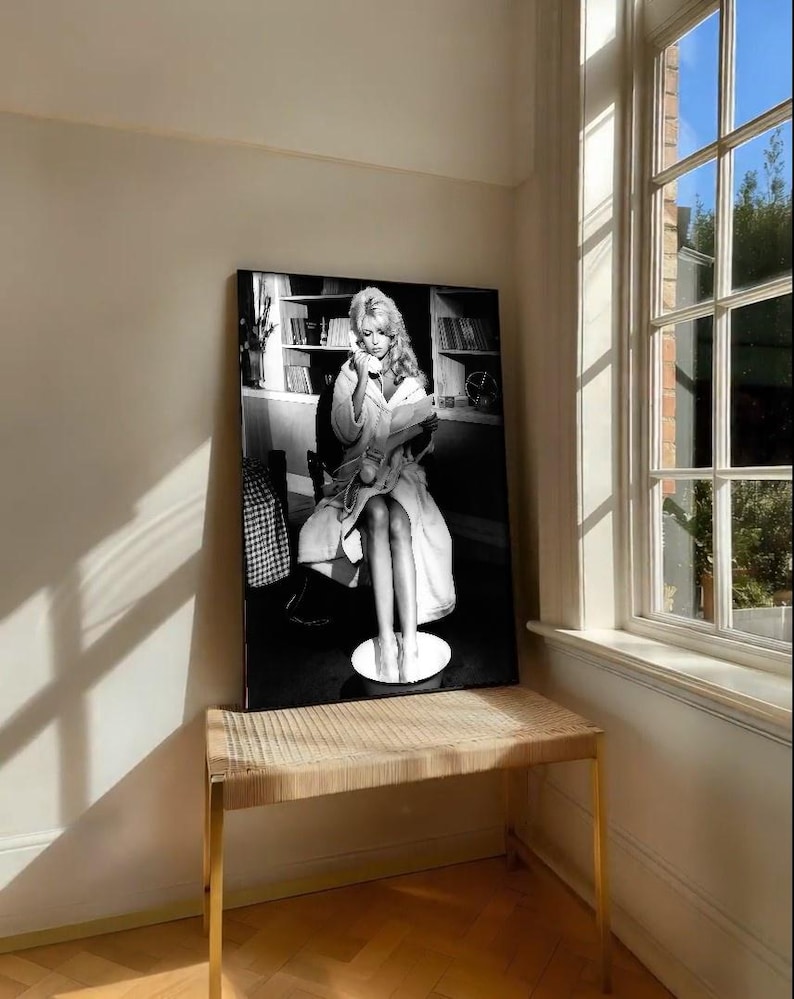 May include: Black and white framed print of a woman in a robe, seated and holding a phone and a book. The print is displayed on a woven bench with gold legs, near a window with natural light.