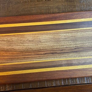 Puede incluir: Una tabla de cortar rectangular de madera con un acabado de madera natural. La tabla tiene tres finas tiras de madera de color dorado incrustadas en la superficie, creando un patrón a rayas.