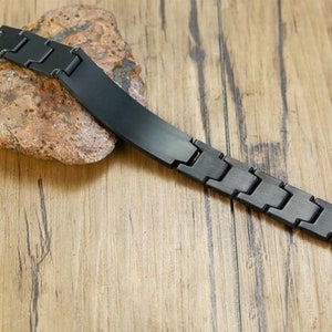 May include: A black metal bracelet with a flat, rectangular plate. The bracelet is made of linked metal pieces.