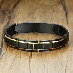 May include: A black and gold stainless steel bracelet with a flat, rectangular plate. The bracelet has a link style design.