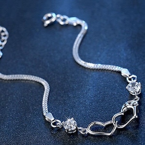 May include: A silver bracelet with a double chain design and three clear crystal accents. The bracelet features two interlocking heart charms.