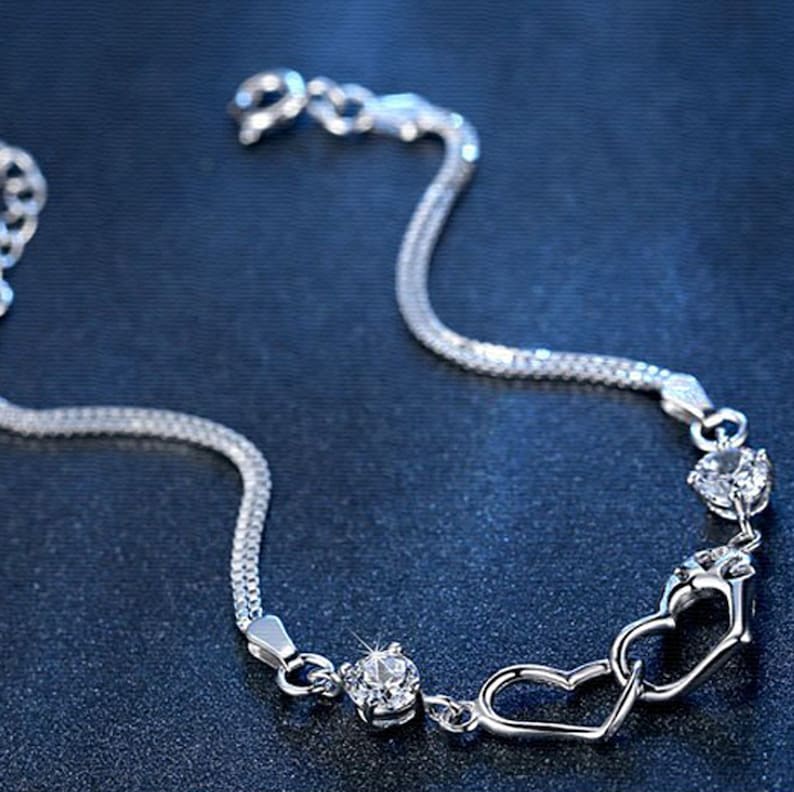 May include: A silver bracelet with a double chain design and three clear crystal accents. The bracelet features two interlocking heart charms.