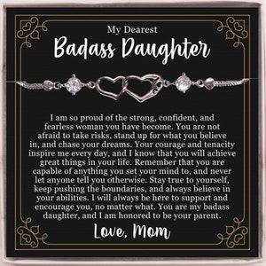 Op de afbeelding: Een zwarte en gouden kaart met een zilveren armband met twee in elkaar verweven harten. De kaart heeft de tekst "Mijn liefste Badass Dochter" en een bemoedigend bericht van een moeder aan haar dochter. De kaart eindigt met de tekst "Liefde, Mama".