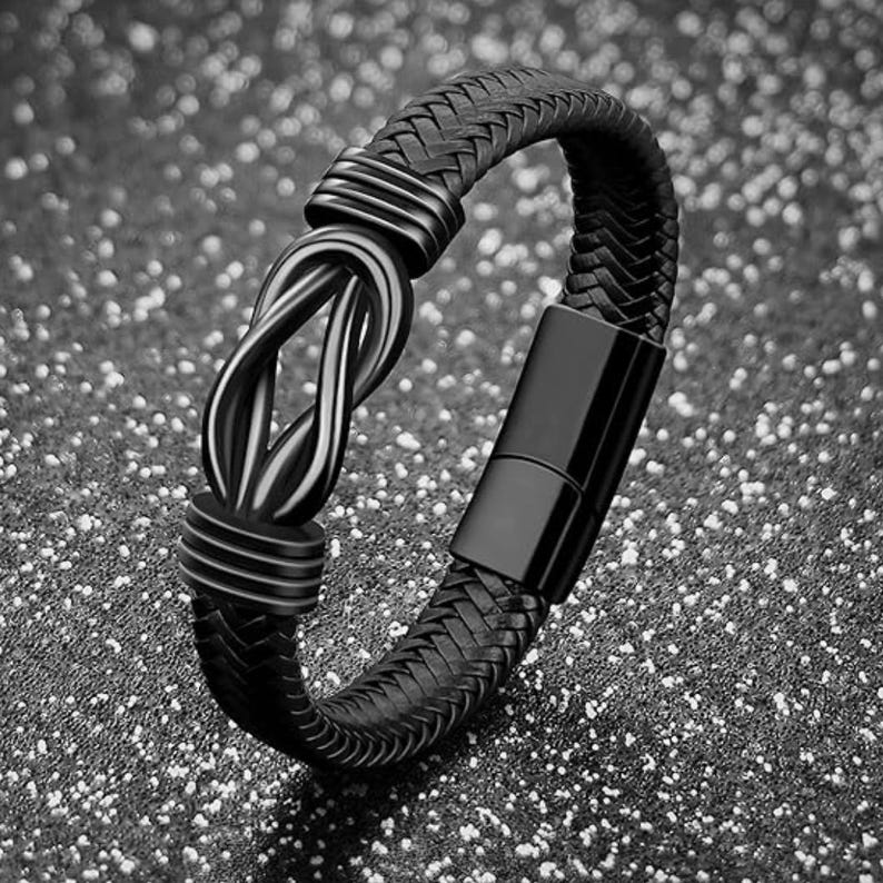 May include: A black leather bracelet with a braided design and a black metal clasp. The bracelet has a knot-like design in the center.