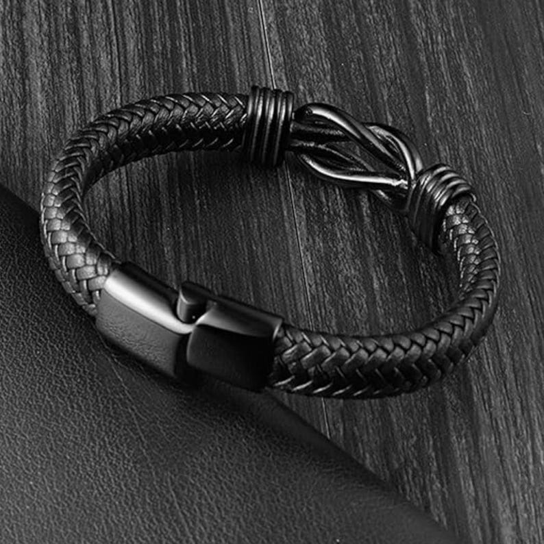May include: Black leather bracelet with a braided design and a black metal clasp. The clasp has a knot-like design.