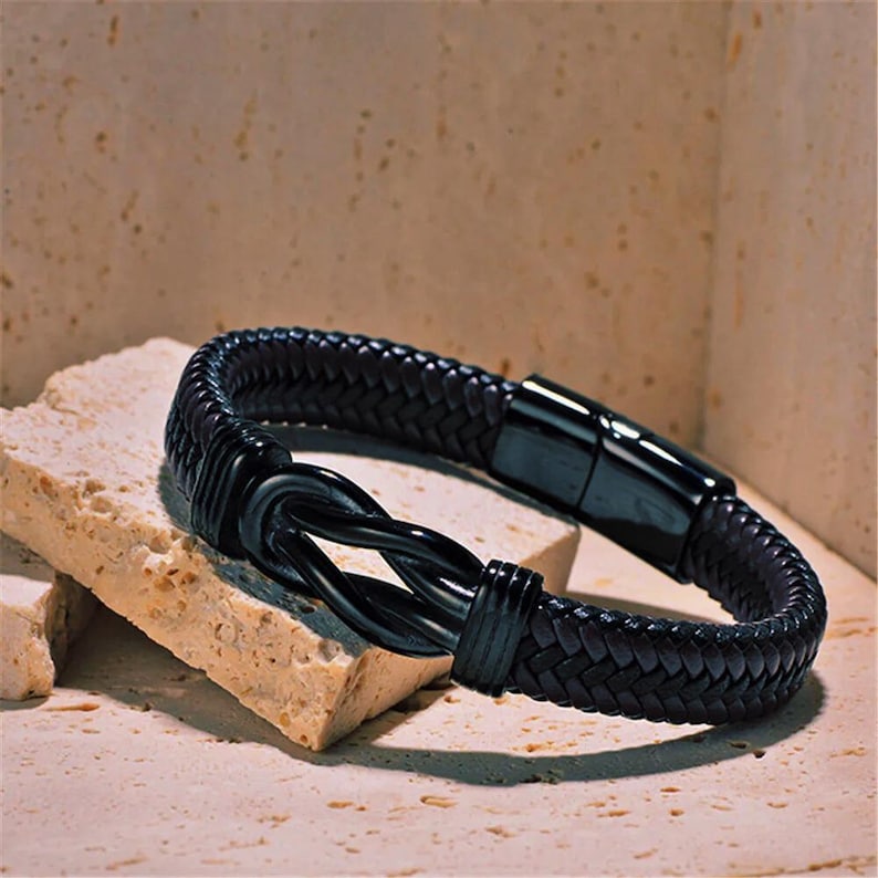 May include: Black leather bracelet with a braided design and a black metal clasp. The clasp features a knot design.