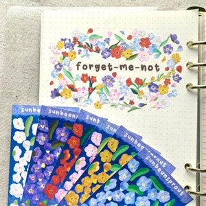 May include: A set of six blue and white floral patterned stickers with the words "sunbeam", "sunbeams", "sunt", "sun", "sunbeam", and "sunbeamsprout" printed on them. The stickers are arranged in a fan shape. The background is a white page with a dotted pattern. The top of the page has a cluster of forget-me-not flowers with the text "forget-me-not" printed in brown.