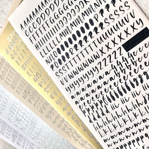 Op de afbeelding: Een set van vijf vellen met alfabetstickers met zwarte letters op witte, gouden en zilveren achtergronden. De letters zijn in een handschrift.