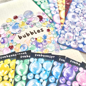 May include: A collection of colorful bubble stickers and sticker sheets. The stickers feature watercolor-style bubbles in various colors, including yellow, blue, purple, and pink. The word "bubbles" is written in brown script. The sticker sheets have the word "sunbeams" on them.