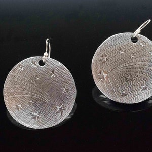 Puede incluir: Un par de pendientes de plata con un diseño redondo y texturado que presenta una estrella fugaz y una dispersión de estrellas.