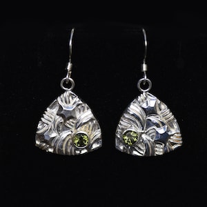 Puede incluir: Un par de pendientes de plata con forma triangular y una piedra preciosa verde en el centro de cada pendiente. Los pendientes tienen una superficie texturizada con un diseño de estrella.