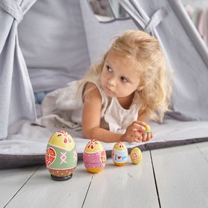 May include: A young girl plays with a set of colourful wooden nesting eggs inside a grey teepee tent. The eggs are decorated with different patterns and designs, including a chicken, hearts, and a floral pattern.