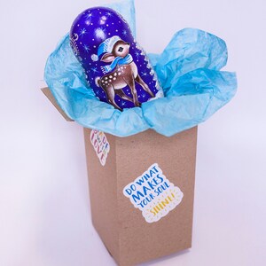 May include: A blue and purple nesting doll with a painted image of a deer wearing a red and white hat. The doll is inside a brown box with a blue tissue paper lining. The box has a sticker that says "Do what makes your soul shine!"