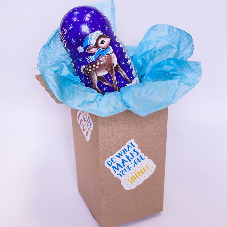 May include: A blue and purple nesting doll with a painted image of a deer wearing a red and white hat. The doll is inside a brown box with a blue tissue paper lining. The box has a sticker that says "Do what makes your soul shine!"