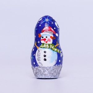 May include: A blue and white hand-painted wooden nesting doll with a snowman design. The snowman is wearing a red hat and a green scarf. The doll is decorated with silver glitter.