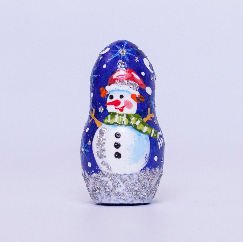 May include: A blue and white hand-painted wooden nesting doll with a snowman design. The snowman is wearing a red hat and a green scarf. The doll is decorated with silver glitter.