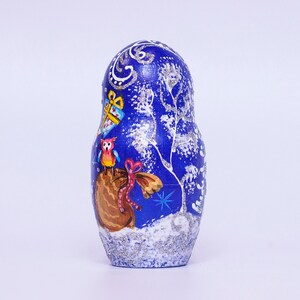 May include: A blue and white hand-painted wooden nesting doll, or matryoshka, with a winter scene. The doll is decorated with a snowy forest, a bird, and a gift box.