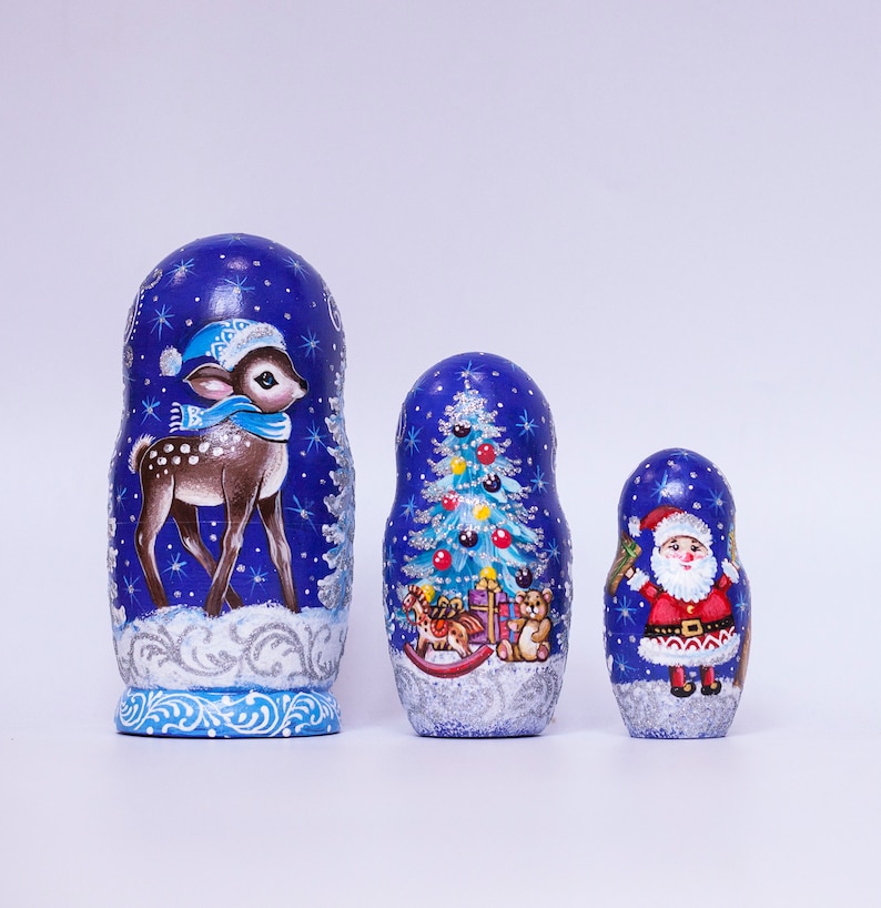 May include: A set of three blue and silver painted Russian nesting dolls, or matryoshka dolls, with winter holiday themes. The largest doll features a deer in a blue hat, the middle doll features a Christmas tree, and the smallest doll features Santa Claus.