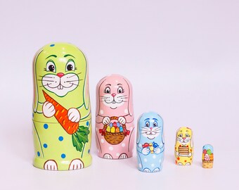 Könnte beinhalten: Satz von fünf Nistpuppen, jede zeigt ein Häschen mit einer anderen Farbe und einem anderen Design. Die größte Puppe ist grün mit weißen Punkten und hält eine Karotte. Die zweitgrößte ist rosa mit weißen Punkten und hält einen Korb mit Eiern. Die drittgrößte ist blau mit weißen Punkten und hält einen Korb mit Süßigkeiten. Die viertgrößte ist gelb mit weißen Punkten und hält einen Kuchen. Die kleinste Puppe ist gelb mit weißen Punkten und hält einen Korb mit Blumen.