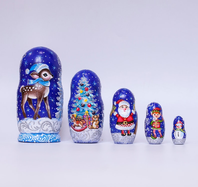 May include: A set of five blue and white painted Russian nesting dolls, or matryoshka dolls, with a winter holiday theme. The largest doll features a deer, the next largest a Christmas tree, the next a Santa Claus, the next an elf, and the smallest a snowman.
