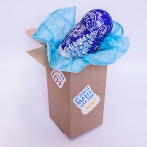 May include: A blue and white hand-painted wooden nesting doll, or matryoshka, is nestled in a brown cardboard box with blue tissue paper. The box has two stickers on it, one that says "Made with Love" and another that says "Do what makes your soul shine!"
