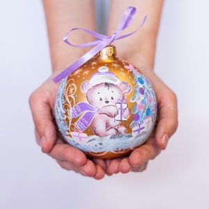 May include: A gold Christmas ornament with a purple ribbon and a painted design of a bear wearing a purple scarf and holding a gift. The bear is surrounded by a blue and white winter scene.