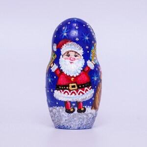 May include: A blue and silver painted wooden nesting doll with a Santa Claus design. The Santa Claus is wearing a red suit with a white beard and a black belt. He is holding a bag of gifts.