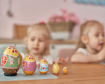 Könnte beinhalten: Ein Set aus sechs bunten, handbemalten Holz-Osterei-Nesting-Puppen. Die Puppen sind in verschiedenen Größen erhältlich, wobei das größte Ei ein grünes und rotes Design aufweist. Das kleinste Ei hat ein gelbes und orangefarbenes Design. Das mittlere Ei hat den Text "I love you".