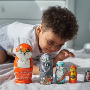 Puede incluir: Un niño mira un conjunto de muñecas rusas de madera de colores, o muñecas matrioska. Las muñecas están pintadas con varios diseños de animales, incluyendo un zorro, un lobo, un gato y una ardilla. Las muñecas están ordenadas por tamaño, con la muñeca más grande a la izquierda y la más pequeña a la derecha.