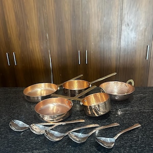 Puede incluir: Una colección de utensilios de cocina de cobre, que incluye cacerolas con mangos largos y una textura martillada, y un juego de cucharas plateadas. Las ollas de cobre tienen una superficie brillante y reflectante. Las cucharas están dispuestas en fila.