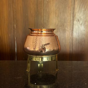May include: A copper drinks dispenser with a hammered finish, complete with a tap and a lid. It is placed on a brass stand with three legs. This dispenser is designed for serving beverages and adds a touch of elegance to any event.