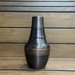 May include: A dark bronze vase with a decorative floral pattern. The vase has a bulbous body and a flared neck. The floral pattern is in a lighter bronze color, creating a contrast against the darker background. The vase is displayed on a wooden surface.