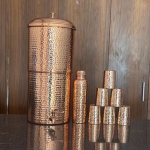 May include: A copper water filter with a hammered texture, a matching bottle, and six copper cups. The water filter has a tap and a lid. The items are arranged on a dark countertop.